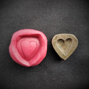 Puede incluir: Dos moldes en forma de corazón, uno de goma roja y el otro de metal. El molde de goma tiene una impresión en forma de corazón en el interior, mientras que el molde de metal tiene un recorte en forma de corazón.