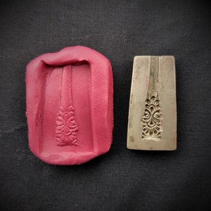 Puede incluir: Un molde de silicona rojo y un sello de metal con un diseño floral. El molde es un rectángulo con un diseño en relieve en el centro. El sello es de forma rectangular con un diseño floral en relieve en el centro.