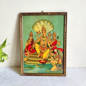 Może przedstawiać: Wizerunek vintage w ramie przedstawiający hinduską boginię Wisznu siedzącego na wielo-głowym wężu z dużym, wielowarstwowym kapturem. Jest otoczony przez trzy inne postacie, wszystkie w ozdobnych nakryciach głowy i tradycyjnych strojach. Wizerunek jest umieszczony na zielonym tle i oprawiony w złotą ramę.