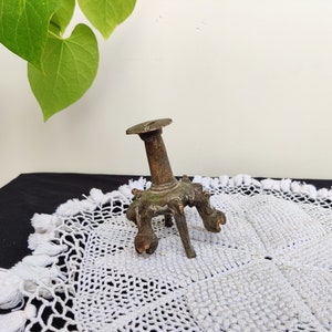 Puede incluir: Un pequeño candelero antiguo de bronce con una parte superior redonda y tres patas ornamentadas. El candelero tiene un aspecto envejecido, con toques de pátina verde. Se encuentra sobre un tapete blanco de ganchillo.