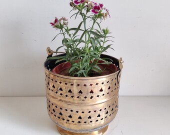 以下が含まれることがあります： ピンクと白の花が咲いた植物が入った、装飾的なカットアウトデザインの真鍮製のプランター