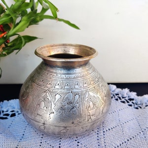 May include: A silver-coloured metal pot with intricate engraved designs, including figures and geometric patterns. The pot has a wide mouth and a rounded body. The pot is sitting on a light blue crocheted surface.