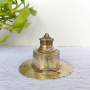 May include: A vintage brass inkwell with a lid. The inkwell has a round base and a tall, cylindrical body. The lid is a dome shape with a small knob on top. The inkwell is tarnished with age, giving it a rustic look.