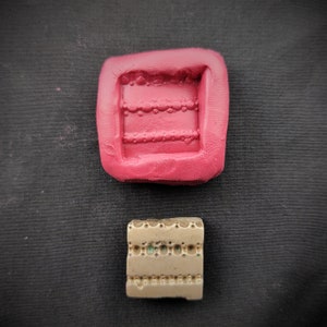 Puede incluir: Dos moldes de silicona para hacer joyas. El molde superior es rosa y tiene un diseño rectangular con un patrón en relieve de círculos y líneas. El molde inferior es marrón y tiene un diseño rectangular similar con un patrón en relieve de círculos y líneas.