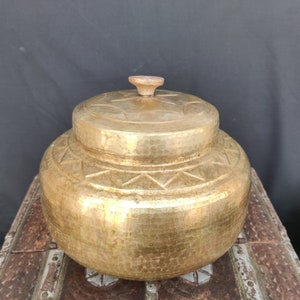 Puede incluir: Un recipiente grande, redondo, de metal de color dorado con una perilla de madera en la tapa. El recipiente tiene una textura martillada y diseños geométricos intrincados alrededor del borde.