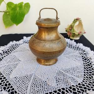 May include: A brass container with a lid and a handle. The container has an ornate design and is sitting on a white crocheted doily.