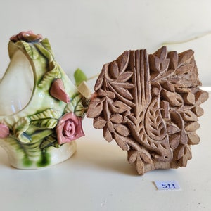 Könnte beinhalten: Zwei Dekorationsartikel, ein Keramiktopf mit Blumenmuster und ein Holzblock mit einem geschnitzten Blumenmuster. Der Topf ist weiß mit grünen und rosa Blumenakzenten. Der Holzblock ist braun und hat ein detailliertes Blumenmuster. Die Nummer 511 ist auf einem weißen Etikett unter dem Holzblock geschrieben.