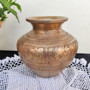 May include: A brass vase with intricate engraved designs of figures and floral motifs. The vase has a rounded body and a flared lip.