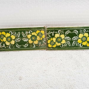 Op de afbeelding: Twee groene en witte keramische tegels met een geel bloemenpatroon. De tegels hebben een rand van groene en witte bladeren.