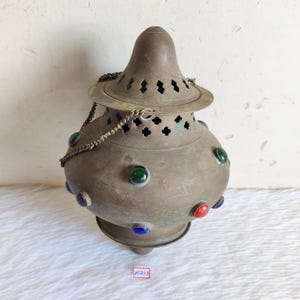 May include: Antique bronze-colored lantern with a conical top and a round body. The lantern features decorative cutouts and is adorned with colorful, round glass accents in red, green, and blue. A chain is attached to the top.