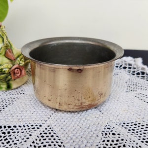 May include: A small, round, brass pot with a slightly tarnished finish. The pot has a smooth, shiny interior and a slightly flared rim.