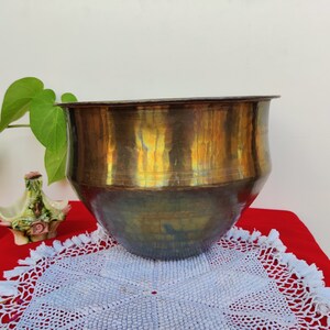 May include: A large, round, brass planter with a hammered finish. The planter has a warm, golden hue and is sitting on a white crocheted doily.