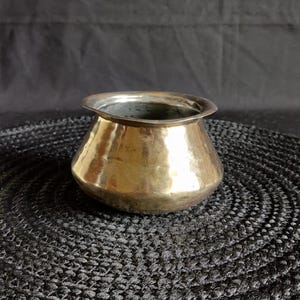 May include: A small, antique brass pot with a rounded body and flared rim. The pot has a polished, reflective surface, and sits on a dark, textured surface. The pot's design is simple and classic, suitable for decorative purposes.