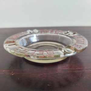 May include: A clear glass ashtray with the words "BEERS" and "CASTLE" printed in red around the rim.