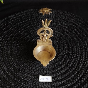 May include: A detailed brass oil lamp with a decorative top and a small bowl-shaped reservoir. The lamp has an ornate design and is placed on a black textured surface. The text "ME 70" is on a small white label.