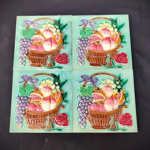 May include: Four square ceramic tiles with a green background and a raised design of a basket of fruit. The fruit includes peaches, pomegranates, and grapes. The basket is brown and has a white handle. The tiles are decorated with flowers.