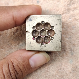 Puede incluir: Un sello de metal plateado con un diseño circular de 12 pequeños círculos dispuestos en forma de flor. El sello se utiliza para crear joyas.