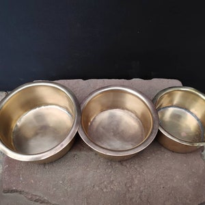 Puede incluir: Tres cuencos de latón de diferentes tamaños, anidados entre sí. Los cuencos son brillantes y tienen una textura ligeramente martillada.