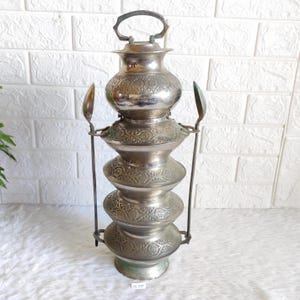 May include: A tall, stacked metal tiffin carrier featuring five rounded, tiered containers with intricate embossed floral patterns. The silver-toned vessel includes a top handle and two vertical side rods, each topped with a spoon-like attachment.