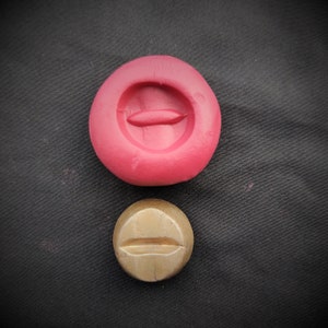 Puede incluir: Dos moldes redondos y planos de resina. El molde más grande es rojo y tiene un diseño ovalado en relieve. El molde más pequeño es dorado y tiene un diseño de línea horizontal en relieve.