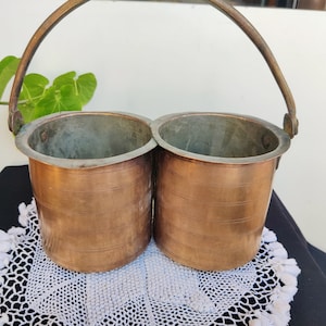 Może przedstawiać: Zestaw dwóch miedzianych wiader z uchwytem, wiadra mają młotkowane wykończenie i stoją na białej koronkowej serwetce.