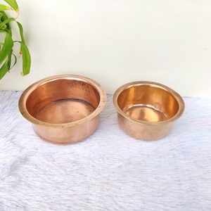 Puede incluir: Dos cuencos de cobre, uno más grande que el otro, se muestran sobre una superficie blanca y peluda. Los cuencos son brillantes y tienen una forma ligeramente redondeada.