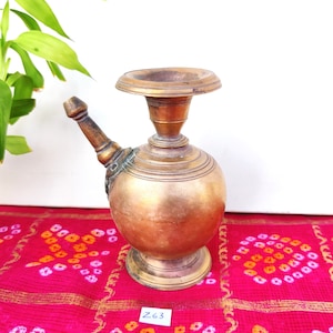 May include: A vintage brass water pot with a spout and a round body. The pot has a decorative design and is sitting on a red and white patterned fabric. The text "Z63" is visible on a white label.