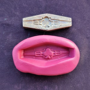 Puede incluir: Un molde de metal plateado y un molde de silicona rosa, ambos con un diseño decorativo con un emblema de trébol. Los moldes tienen forma de rectángulo estilizado con bordes redondeados. El molde de metal está detallado con remaches.