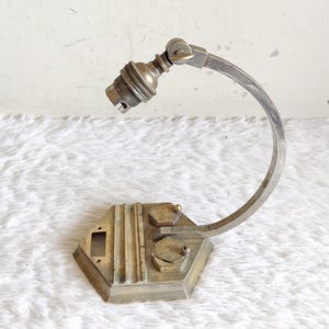May include: An antique brass-toned desk lamp with a curved arm and a hexagonal base. The lamp features a bulb socket and a pivoting head. The base has a rectangular cutout and decorative ridges.
