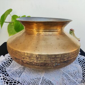 May include: A large, round, brass pot with a wide, flared rim. The pot has a worn, aged patina and is sitting on a white lace doily.