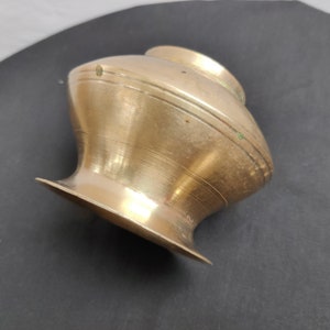 Puede incluir: Una pequeña olla de latón antiguo con base acampanada y cuerpo redondeado. La olla tiene un cuello estrecho y un borde ligeramente más ancho. El latón tiene un color dorado cálido con algunos signos de edad y desgaste. La olla probablemente se utiliza con fines decorativos o ceremoniales.