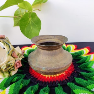 Puede incluir: Una olla de latón vintage con una base ancha y redonda y un borde ligeramente acampanado. La olla está sentada sobre un encaje de ganchillo colorido con acentos verdes, rojos, amarillos y blancos.