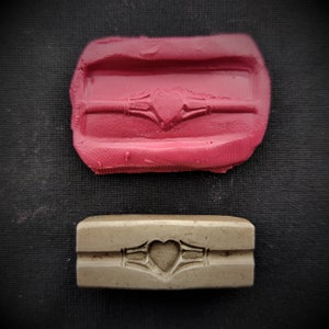 Puede incluir: Dos moldes rectangulares para hacer joyas. El molde superior es rosa y el molde inferior es marrón. Ambos moldes tienen un diseño de corazón con un patrón de nudo celta.