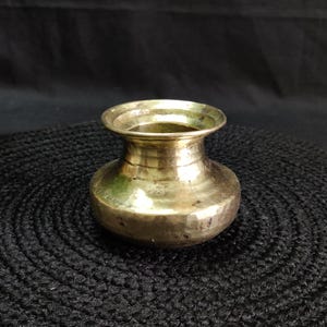 May include: A small, antique brass pot with a wide, rounded body and a flared rim. The pot has a hammered texture and a tarnished, aged appearance. The pot is likely used for decorative purposes.