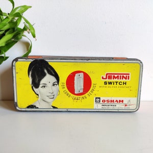 Può includere: Una scatola di latta vintage con sfondo giallo e un viso di donna sul davanti. La scatola pubblicizza "Jemini Switch with Silver Contact" e "Osham Industries." Il testo "For Long-Lasting Service" è anche sulla scatola.