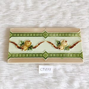 May include: A vintage ceramic tile with a green and white border and a floral design featuring yellow flowers and brown vines. The tile has a beige background and a diamond pattern in the corners.