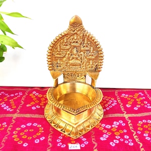 May include: A gold metal diya with intricate carvings of deities. The diya has a heart-shaped base and a raised back with a central figure surrounded by other figures. The diya is ideal for traditional Indian ceremonies and celebrations.