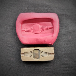 Puede incluir: Un molde de silicona rosa y un objeto metálico rectangular. El molde es de color rosa brillante y tiene una impresión detallada del objeto metálico. El objeto metálico es rectangular con un diseño central y líneas decorativas.