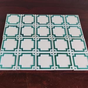 May include: A collection of sixteen white tiles with a green geometric border design. The tiles are arranged in a rectangular pattern on a dark brown surface. The design features a square within a square.