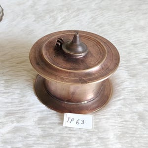 May include: A vintage, copper-toned metal container with a lid and a small handle. The container has a circular base and a tiered design. The lid features a conical knob. The label on the bottom right reads "IP 63".