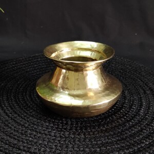 Puede incluir: Una pequeña olla de latón martillado con un borde ancho y acampanado y un cuerpo redondeado. La olla tiene un acabado dorado brillante y se asienta sobre una superficie negra texturizada. El diseño de la olla sugiere que podría usarse con fines decorativos.