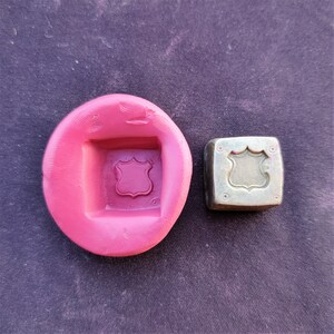 Puede incluir: Un molde de silicona rosa con una impresión cuadrada de un diseño de escudo. Junto al molde hay un sello de metal con el mismo diseño de escudo.