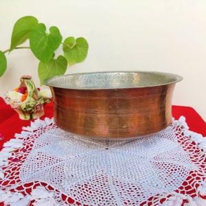 May include: A large, round copper bowl with a hammered finish sits on a white crocheted doily. The bowl is on a red surface.