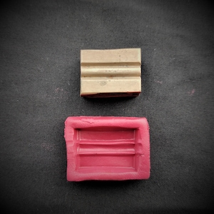 Puede incluir: Dos moldes de jabón rectangulares, uno es beige y el otro es rosa. Ambos moldes tienen tres líneas elevadas en el interior.