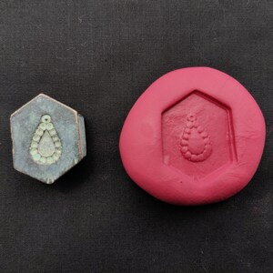 May include: A hexagonal mould for making jewellery, with a teardrop-shaped design, is shown next to a metal stamp with the same design. The mould is made of pink silicone and the stamp is made of metal with a green patina.