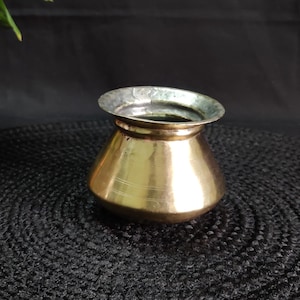 Puede incluir: Una pequeña olla de latón antiguo con un borde plateado. La olla tiene un cuerpo redondeado con líneas horizontales y una abertura ligeramente ensanchada. El fondo es oscuro, resaltando los tonos cálidos de la olla.