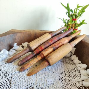 May include: Four wooden rolling pins, two with red and white stripes, in a wooden bowl with a white crocheted doily. The rolling pins are arranged in a fan shape. The bowl is on a white surface. E76 is written on a white tag.