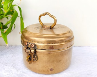 Puede incluir: Un pequeño recipiente redondo de latón con tapa con bisagras y asa decorativa. El recipiente tiene un aspecto desgastado y antiguo.