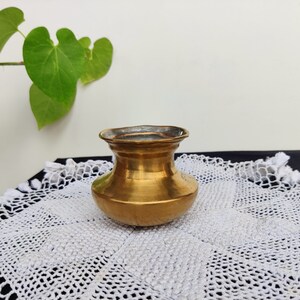 Puede incluir: Una pequeña olla de latón antiguo con cuerpo redondeado y borde acampanado. La olla tiene un tono dorado y se asienta sobre un mantelito blanco y de encaje. Se ven hojas verdes en el fondo. Esta olla decorativa es ideal para la decoración del hogar.