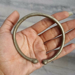 Könnte beinhalten: Ein silberfarbenes, offenes Armband mit leicht strukturierter Oberfläche. Das Armband hat ein kreisförmiges Design mit geometrischen Endkappen. Das Armband wird in einer offenen Hand gehalten.